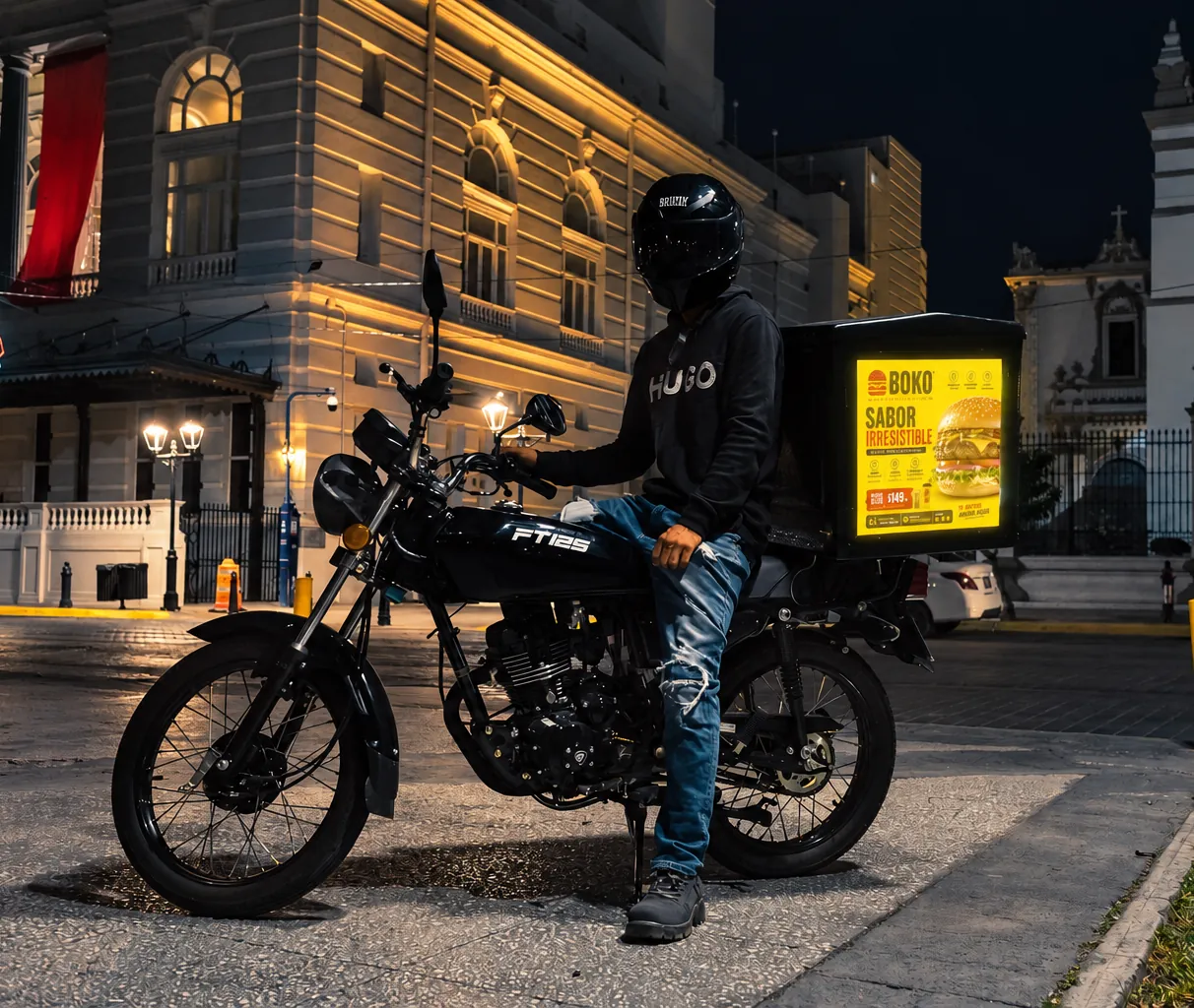 Repartidor sentado en su moto de noche, con la caja Urbanet encendida